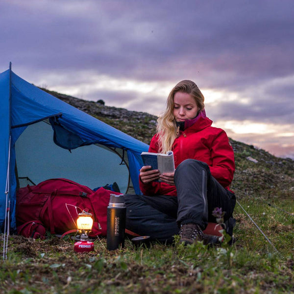 Lifestyle shot outdoors of a seated woman reading a book by the light of a Primus Easy Light lantern outside her tent